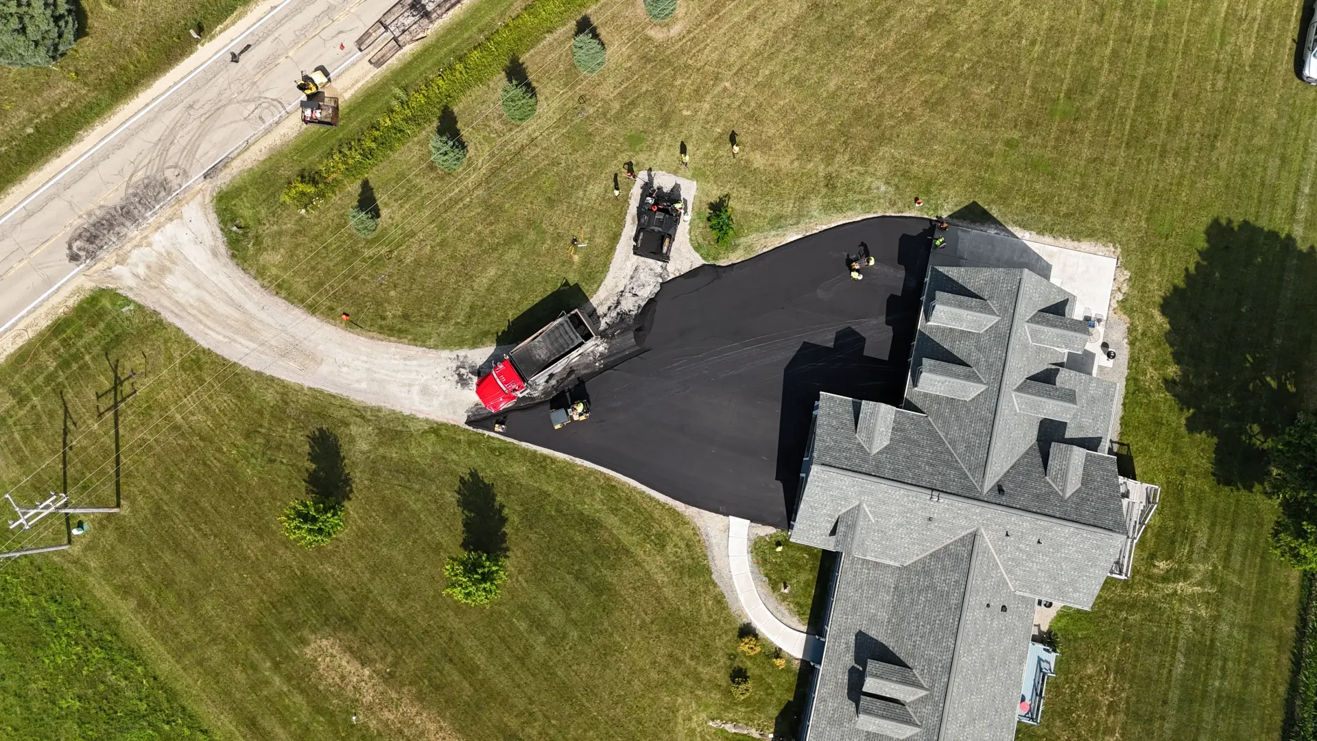 Aerial view of a house with a red car parked on a dark driveway beside a curved gravel road.