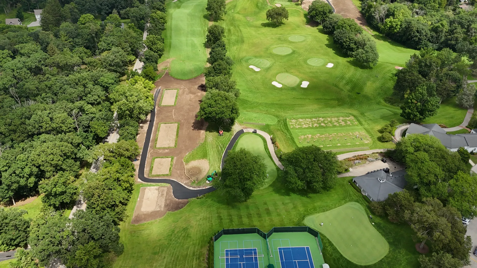 Aerial view of a golf course with fairways, sand traps, trees, and blue tennis courts at the bottom.