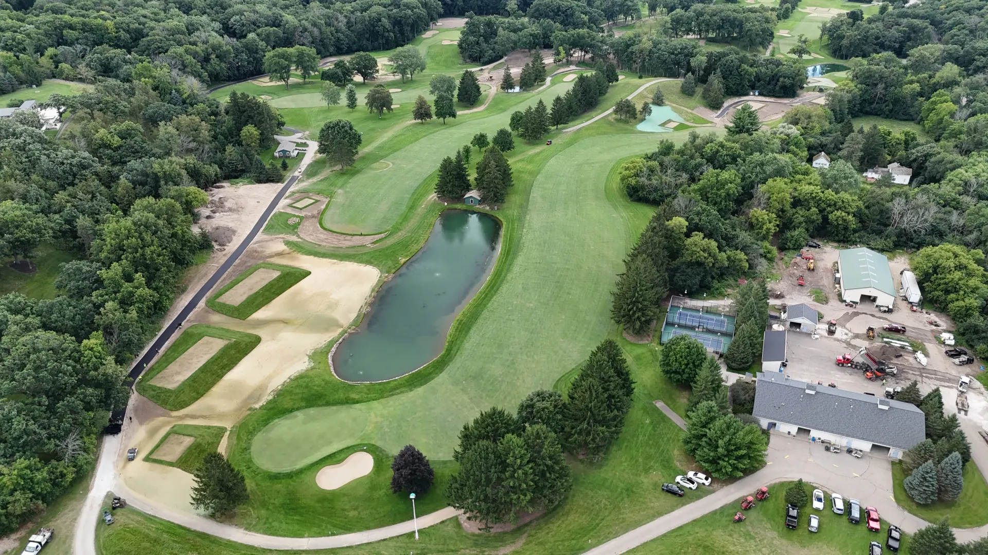 Aerial view of a golf course with fairways, sand bunkers, a water hazard, and nearby buildings.