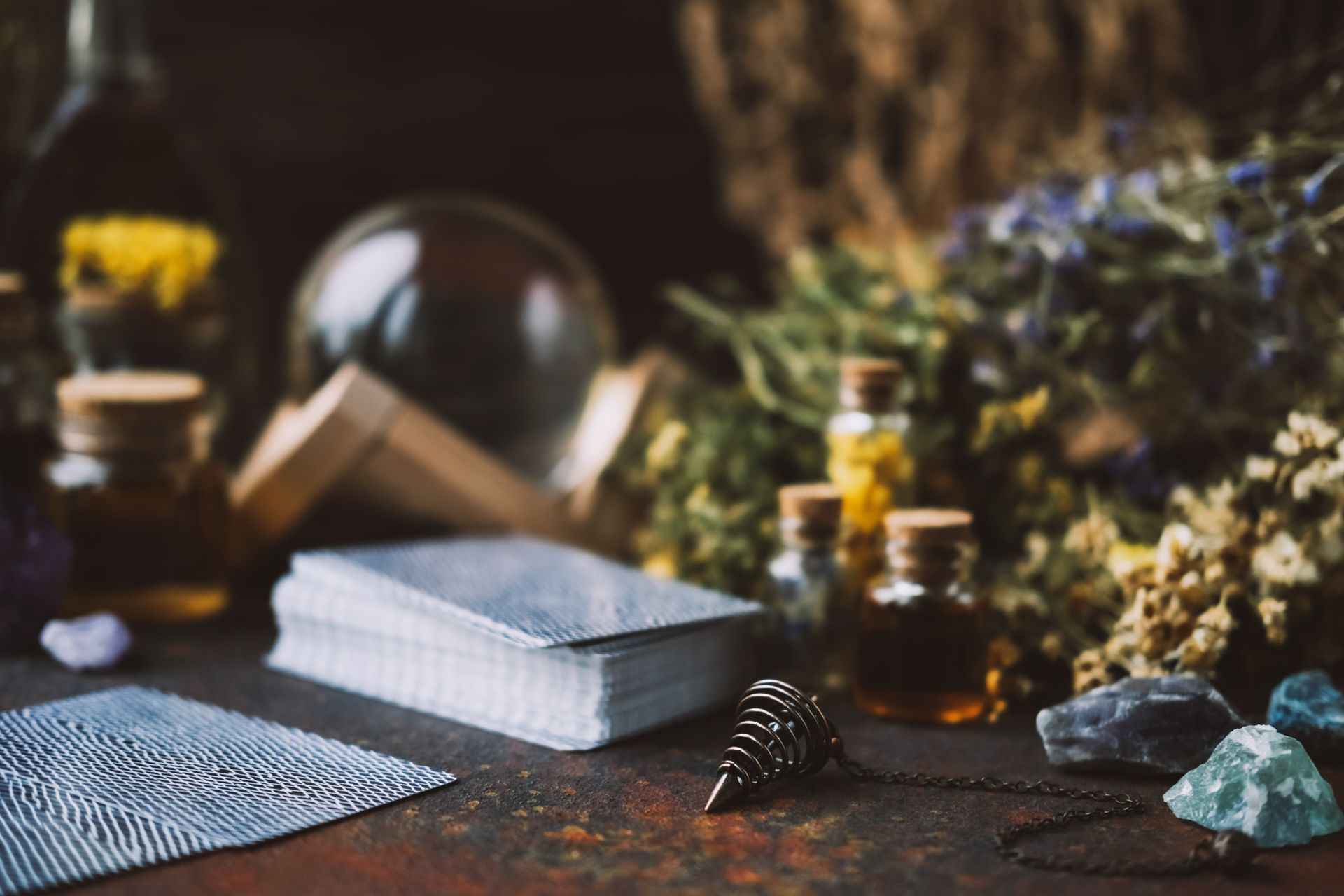 A pendulum, tarot cards, and stones on a table against blurred flowers, glass jars, & a crystal ball.