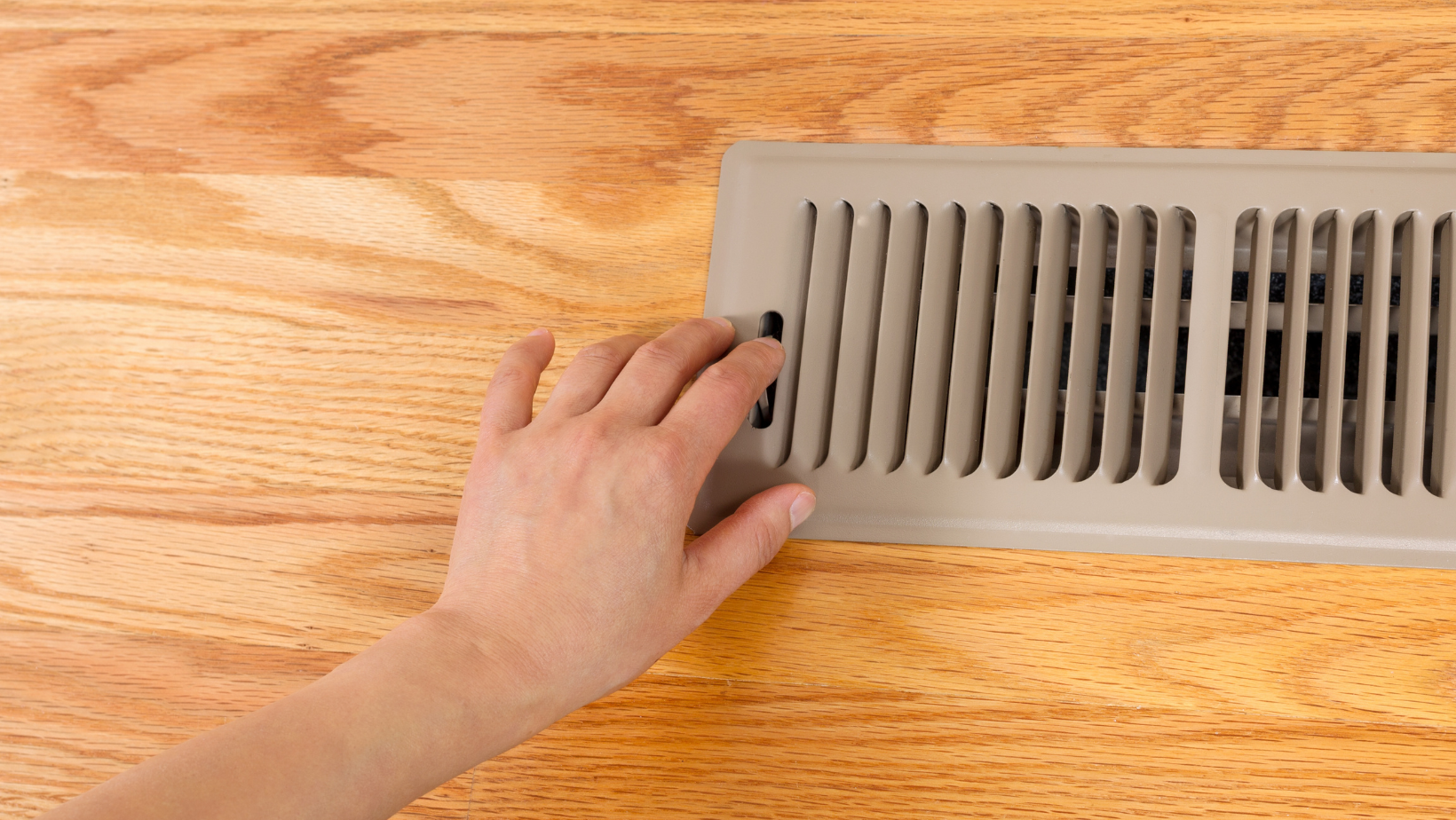Hand reaching towards a floor vent on a wooden floor.