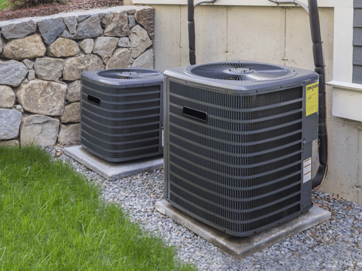 Two outdoor air conditioning units on concrete pads beside a building and gravel.