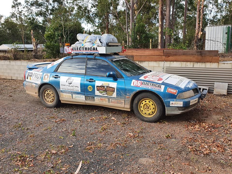 Blue rally car with roof cargo and sponsor decals parked on a gravel lot beside trees and a wall