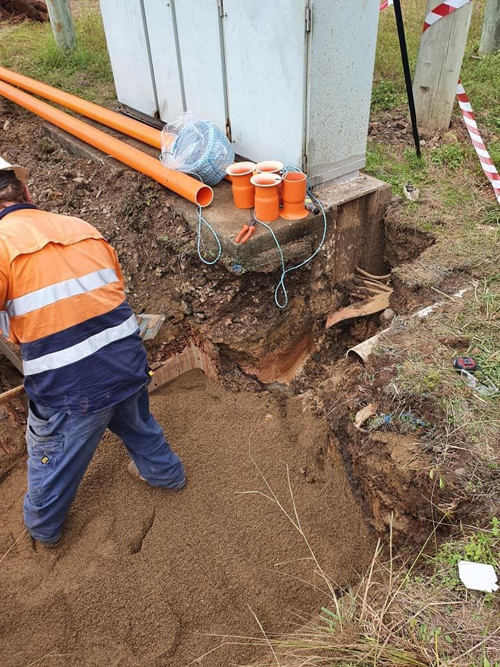 Worker in orange vest near a muddy excavation with orange pipes and utility boxes beside a wall