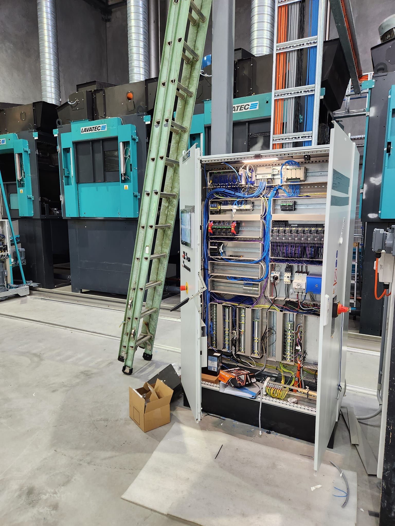 Industrial electrical cabinet open in a workshop, with exposed wiring, ladders, and machinery in the background.