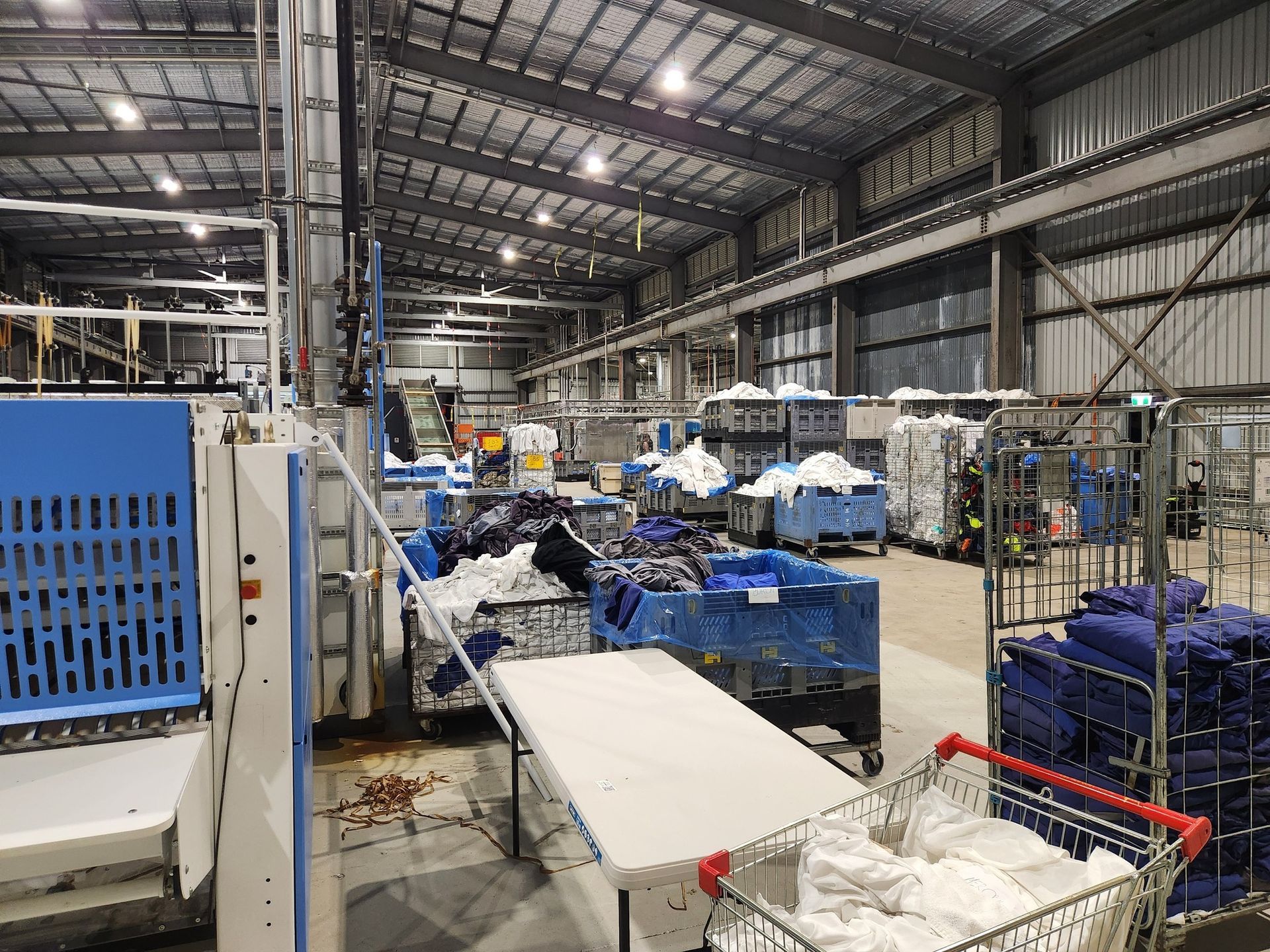 Inside a warehouse, blue bins and stacked materials surround a conveyor line under industrial lighting.