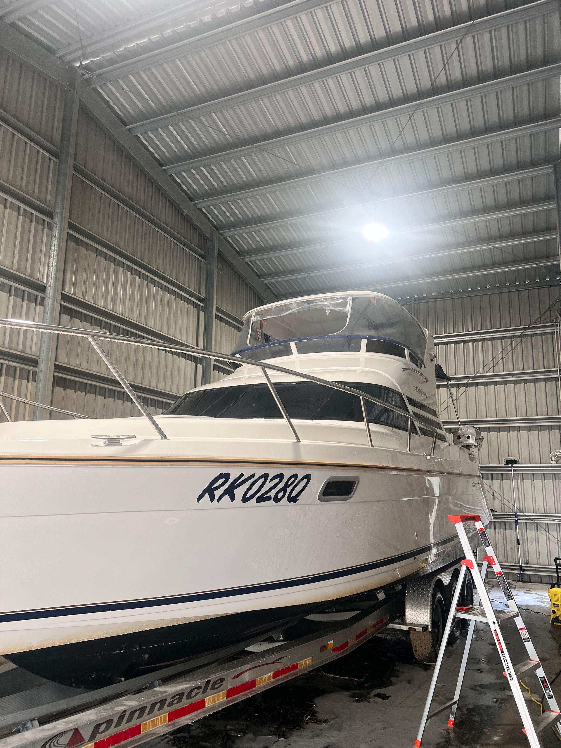 A white motorboat with the registration RK028Q sits on a trailer inside a large, metal-walled storage shed.