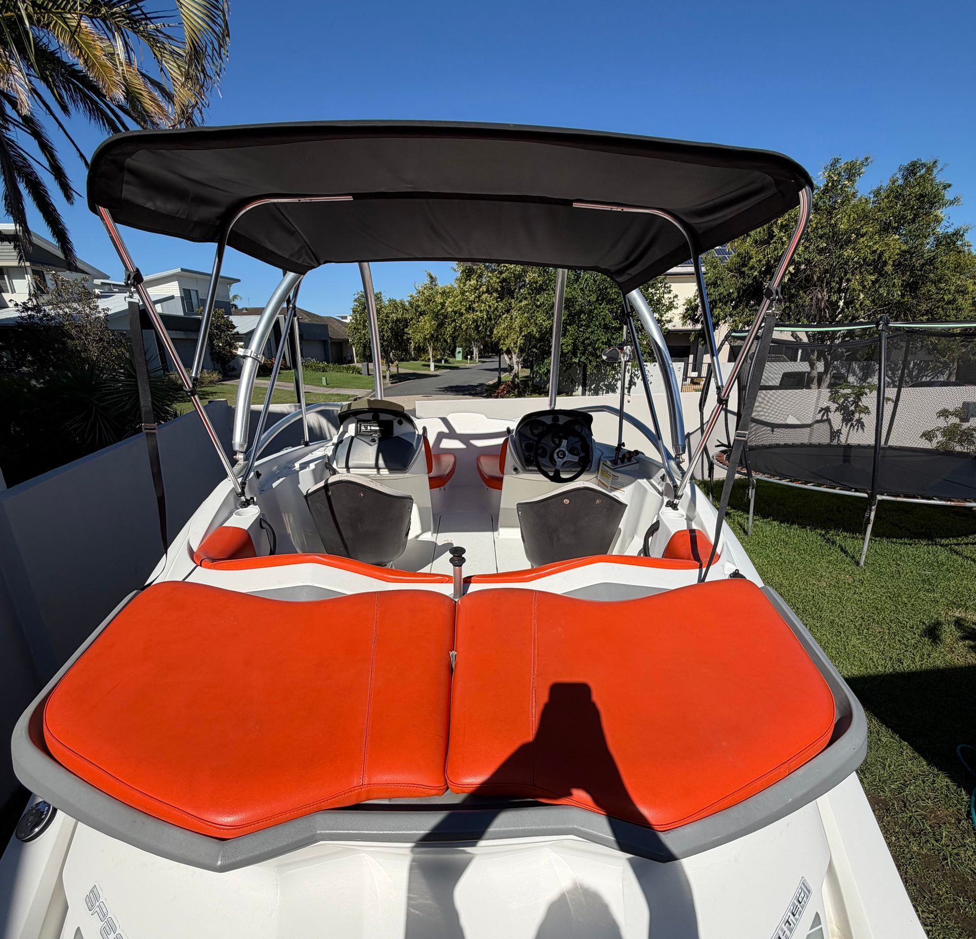 Rear view of a boat featuring bright orange sun pads, a black bimini top, and a white deck, parked outdoors on grass.