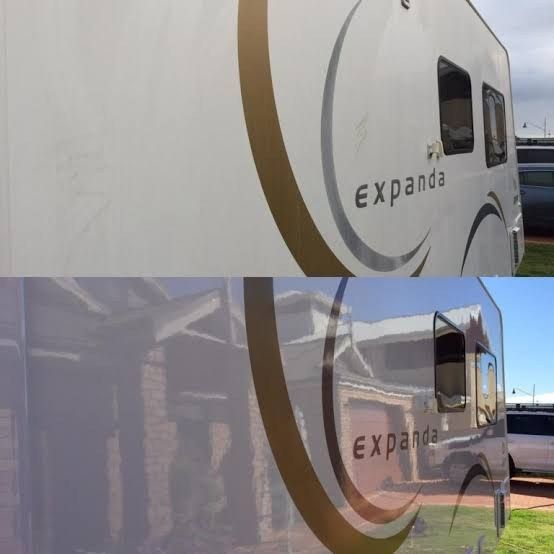 A beige travel trailer with orange, black, and grey graphic accents parked on a paved lot with trees in the background.