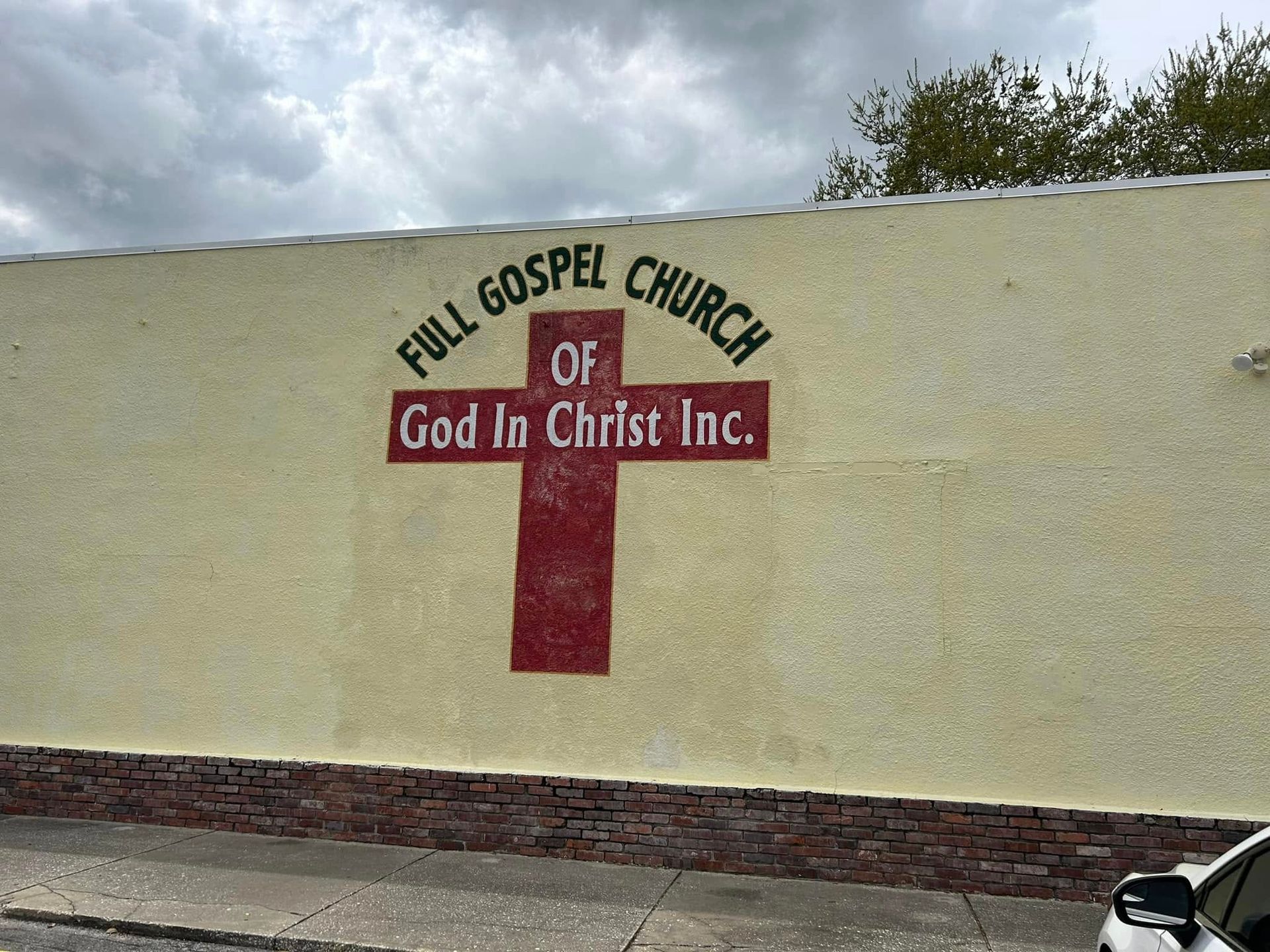 Full Gospel Church of God In Christ Inc. sign on cream wall with red cross, brick base, cloudy sky.
