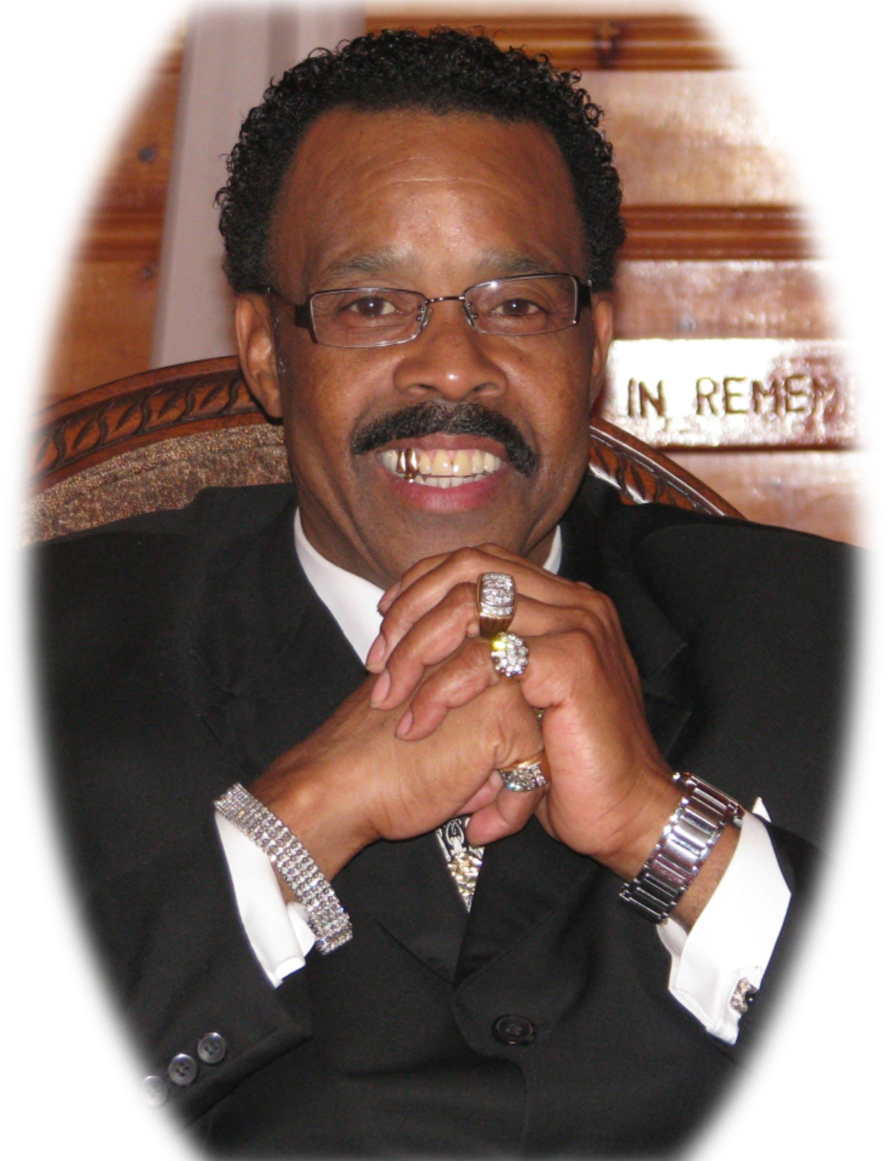 Man in black suit, smiling with hands clasped, wearing glasses and jewelry.