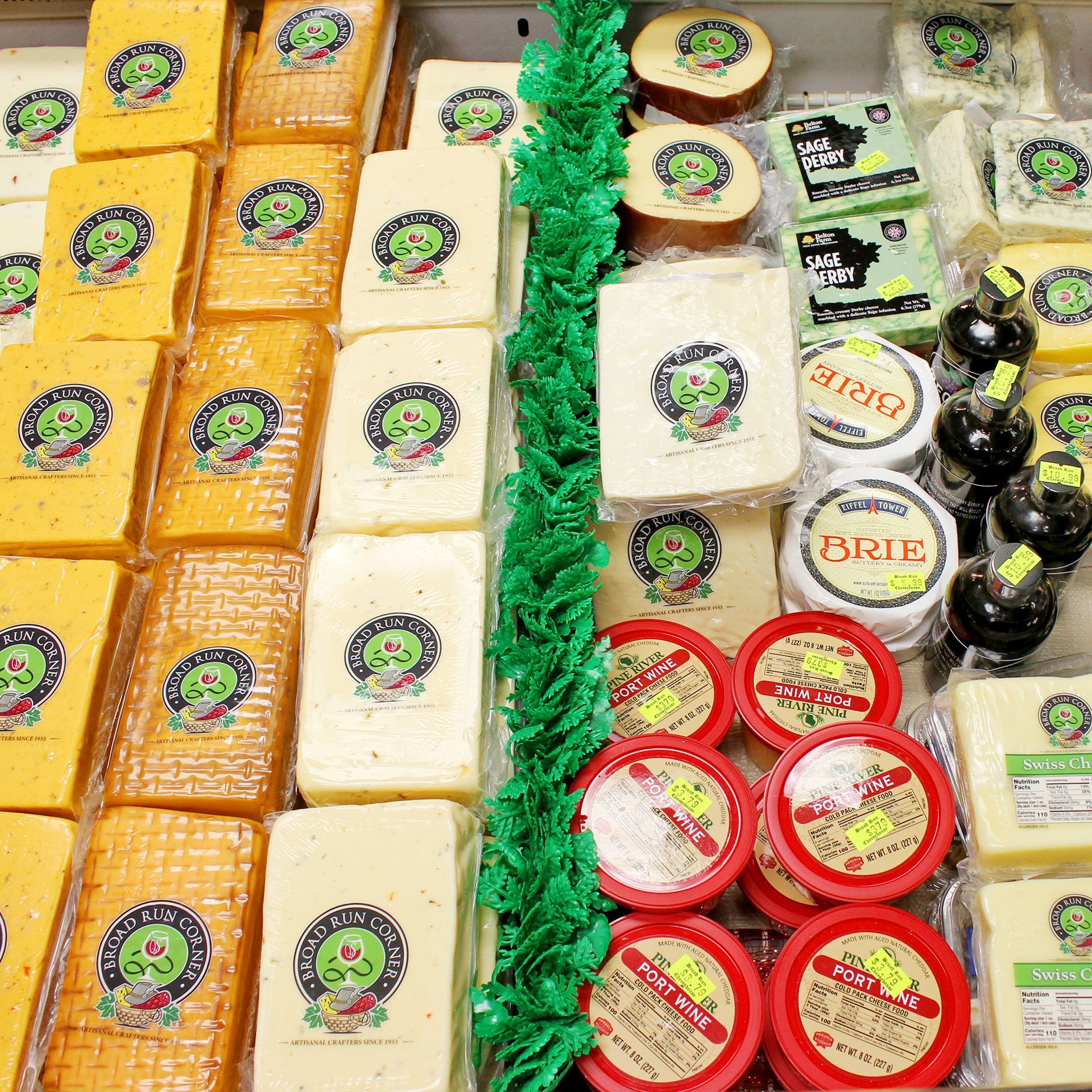 Various cheeses in a refrigerated display case. Yellow, white, and red cheeses are arranged with green decorative accents.