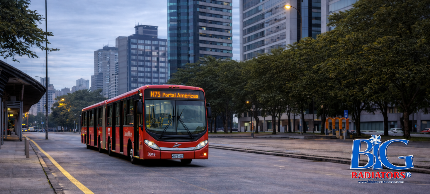 Autobús articulado rojo circulando por una calle de la ciudad al anochecer, con edificios altos y árboles al fondo.