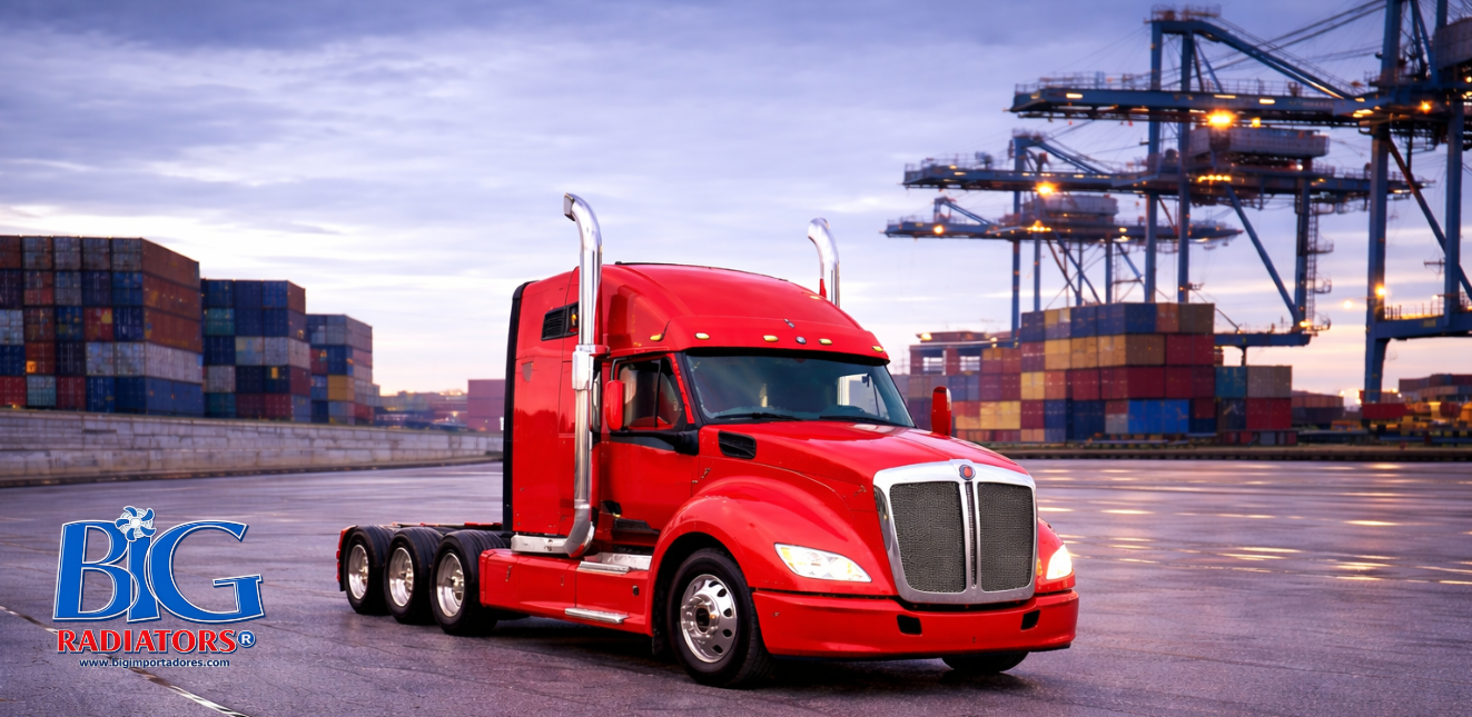 Un camión semirremolque rojo circula por una terminal de carga con contenedores apilados y grúas.