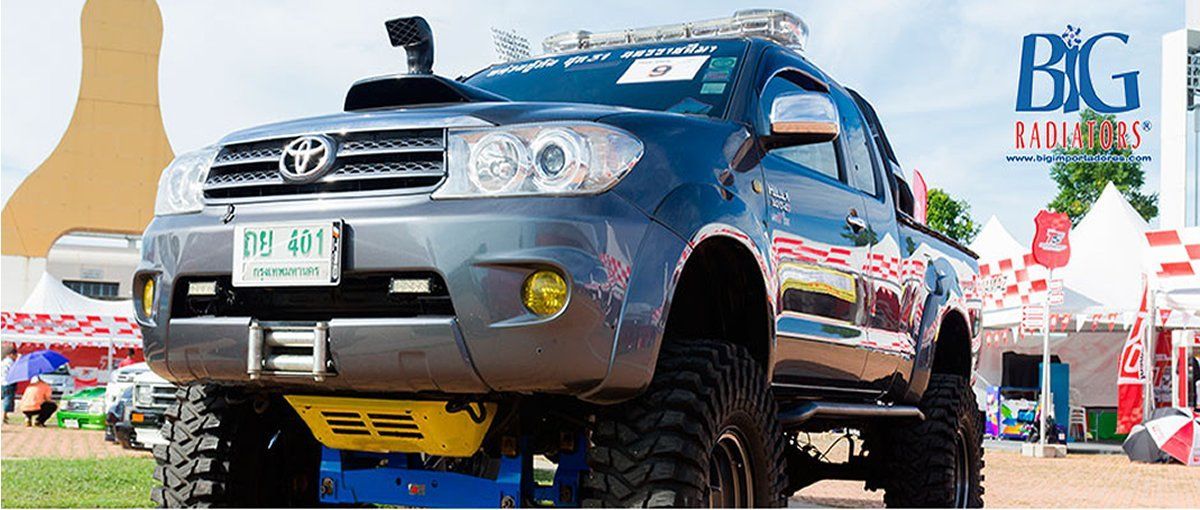 SUV azul todoterreno en un elevador en un evento al aire libre, con carpas y un logotipo grande al fondo.