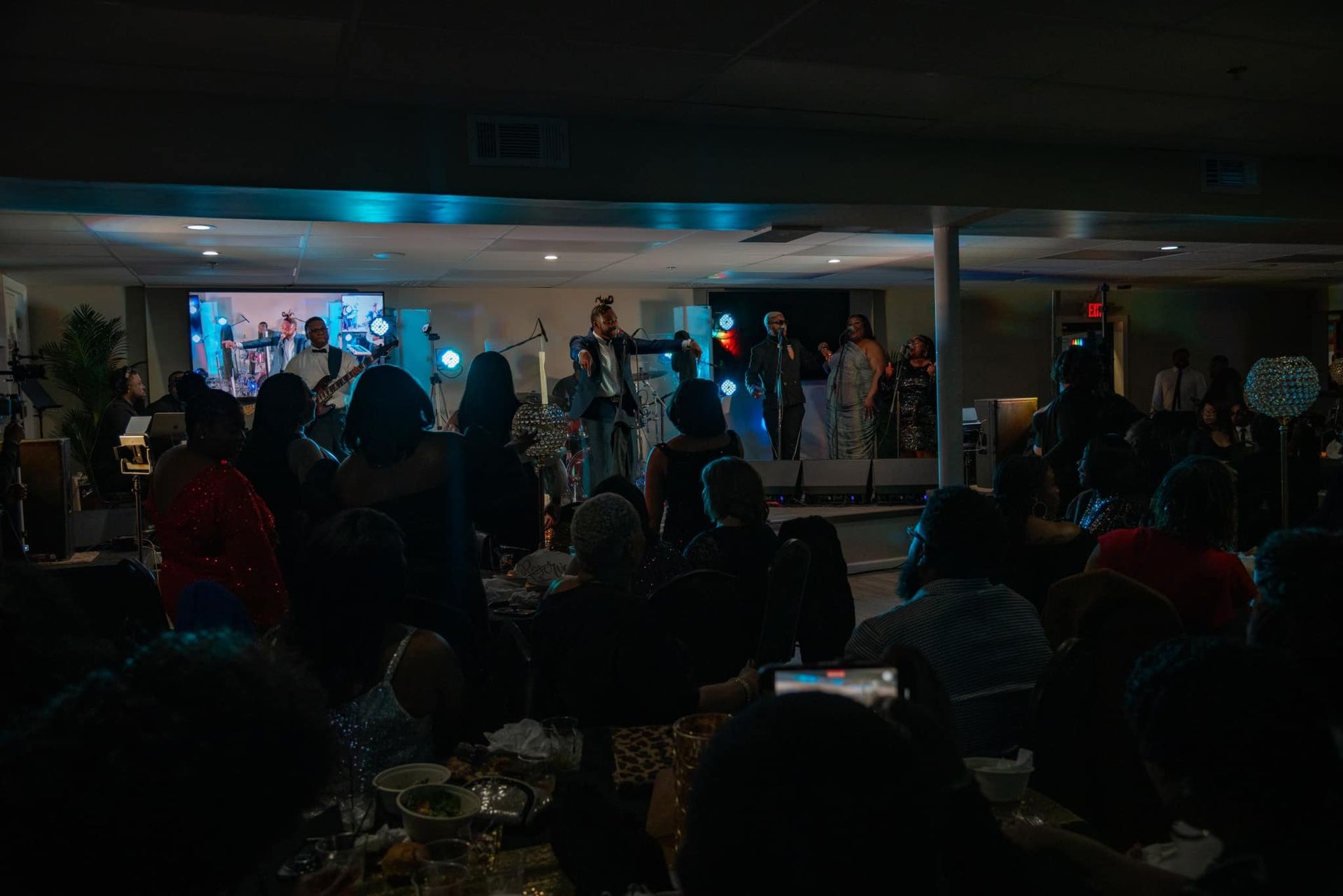 A group of people are sitting at tables in a dark room watching a band perform.