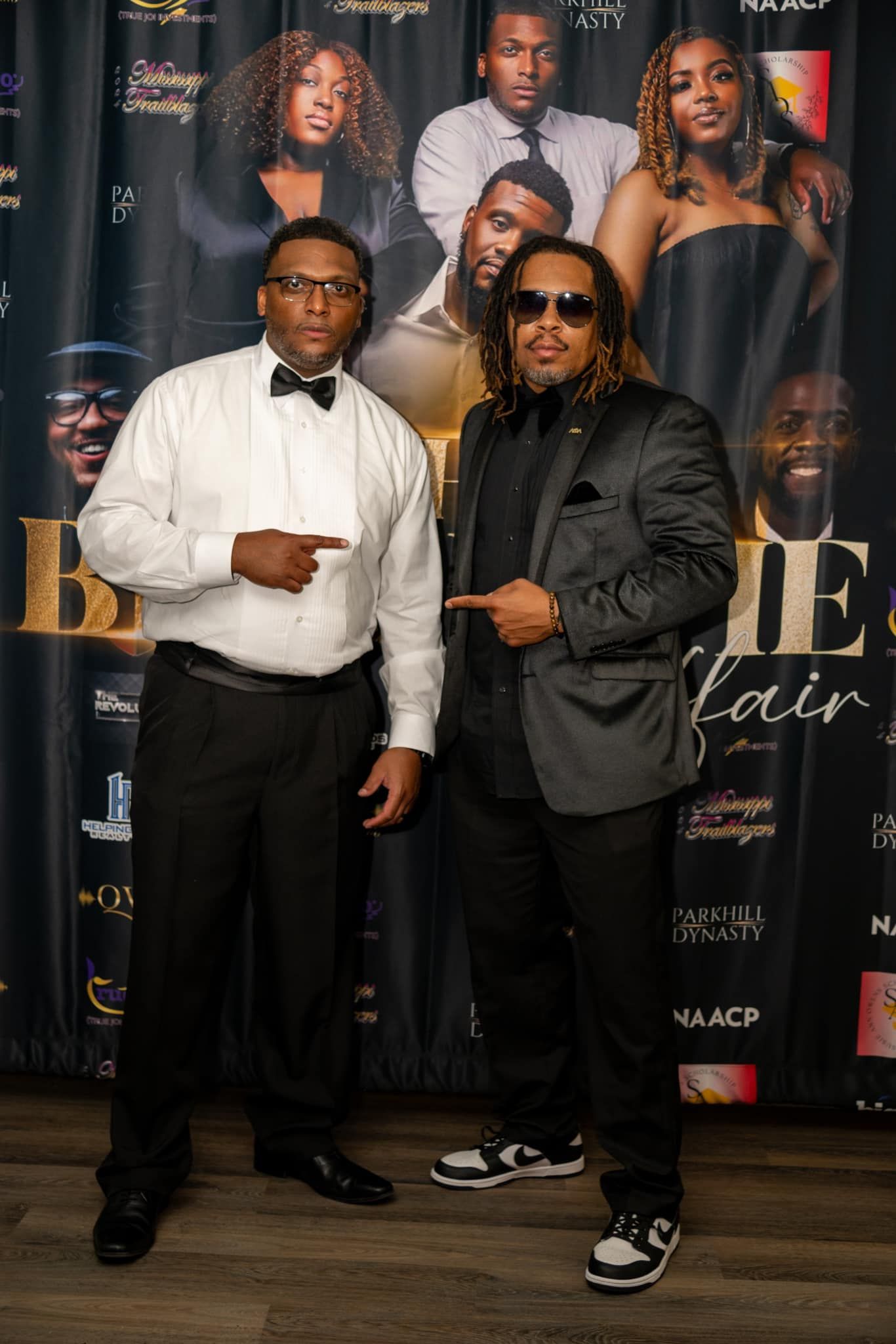 Two men in suits are standing next to each other in front of a black curtain.