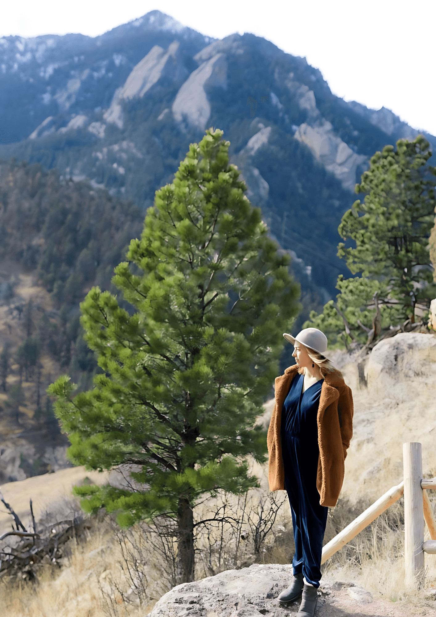 Woman in brown coat and hat stands on a rocky overlook, mountains in the background.