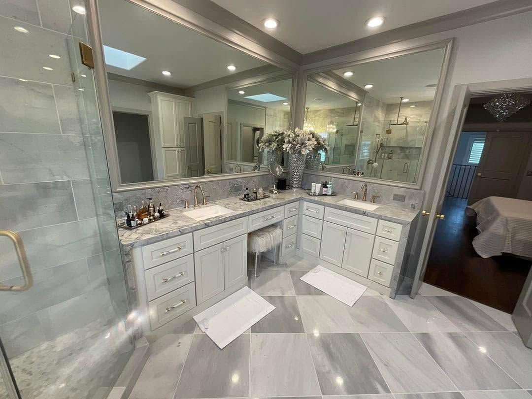 Bright white marble bathroom with double vanity, large mirror, glass shower, and gray floor tiles.