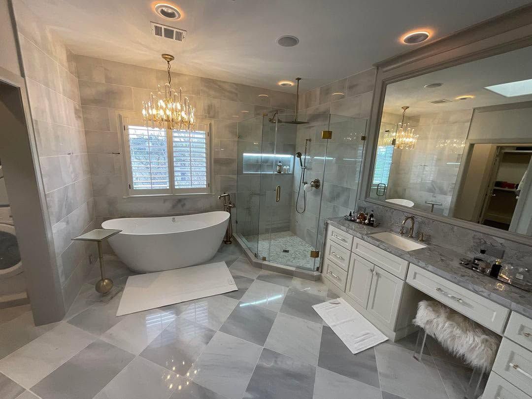 Elegant white bathroom with chandelier, freestanding tub, glass shower, and large vanity mirror.