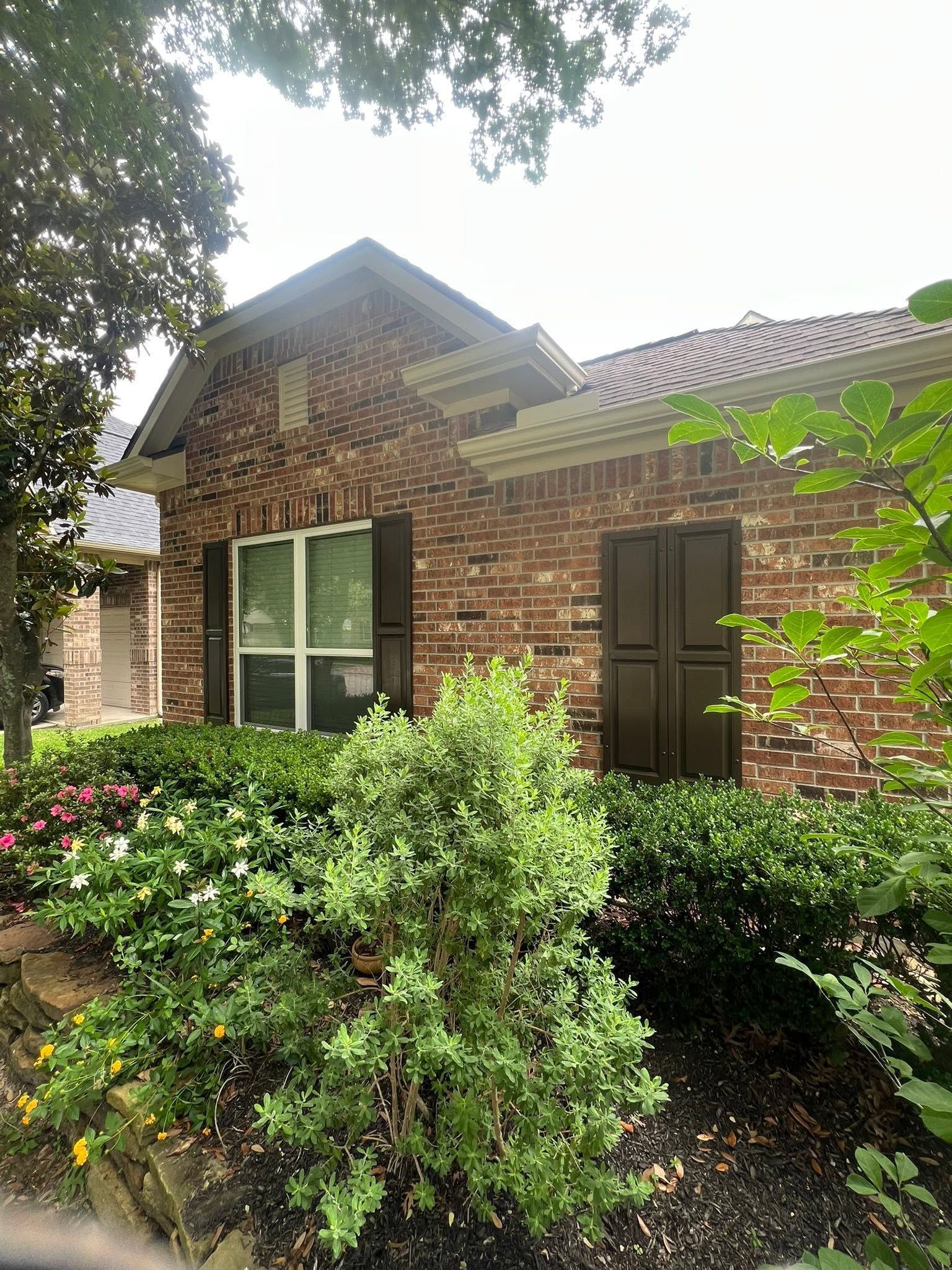 Brick house front with green shrubs, white-trimmed window, and brown door under trees