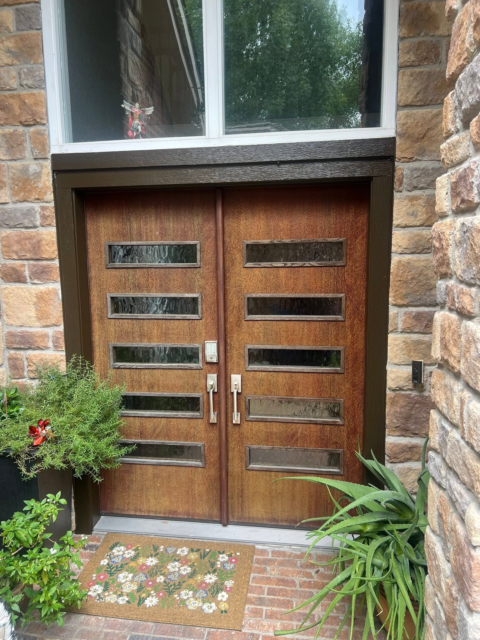 Wooden double front doors with horizontal glass slits, framed by brick and greenery.
