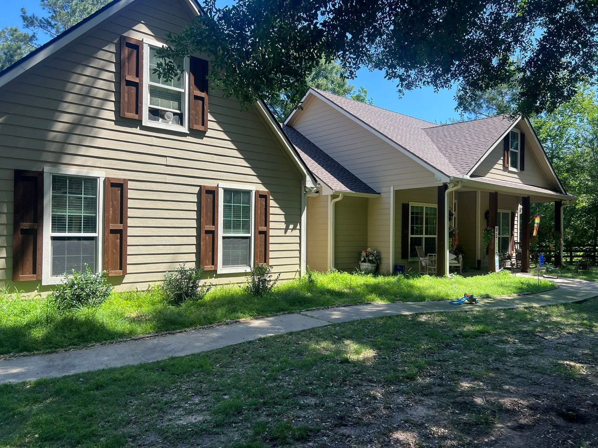 Two-story tan house with brown shutters and a covered front porch, surrounded by trees and grass.