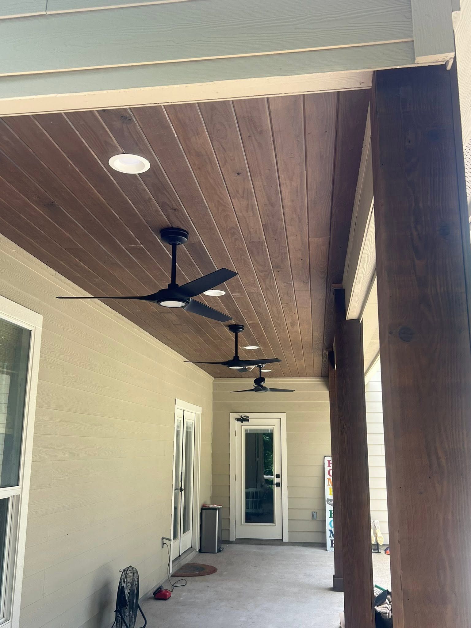 Covered porch with wood ceiling, black ceiling fans, and cream walls beside a doorway