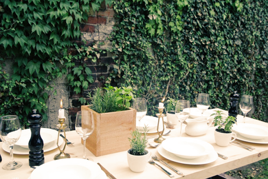 Outdoor table set with white tableware, candles, and greenery against a vine-covered brick wall