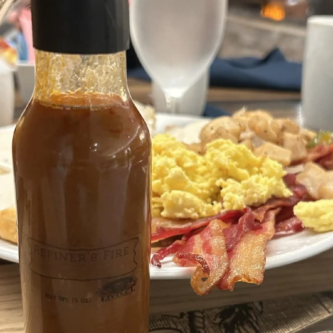 A bottle of hot sauce in front of a plate of breakfast with eggs, bacon, and potatoes.