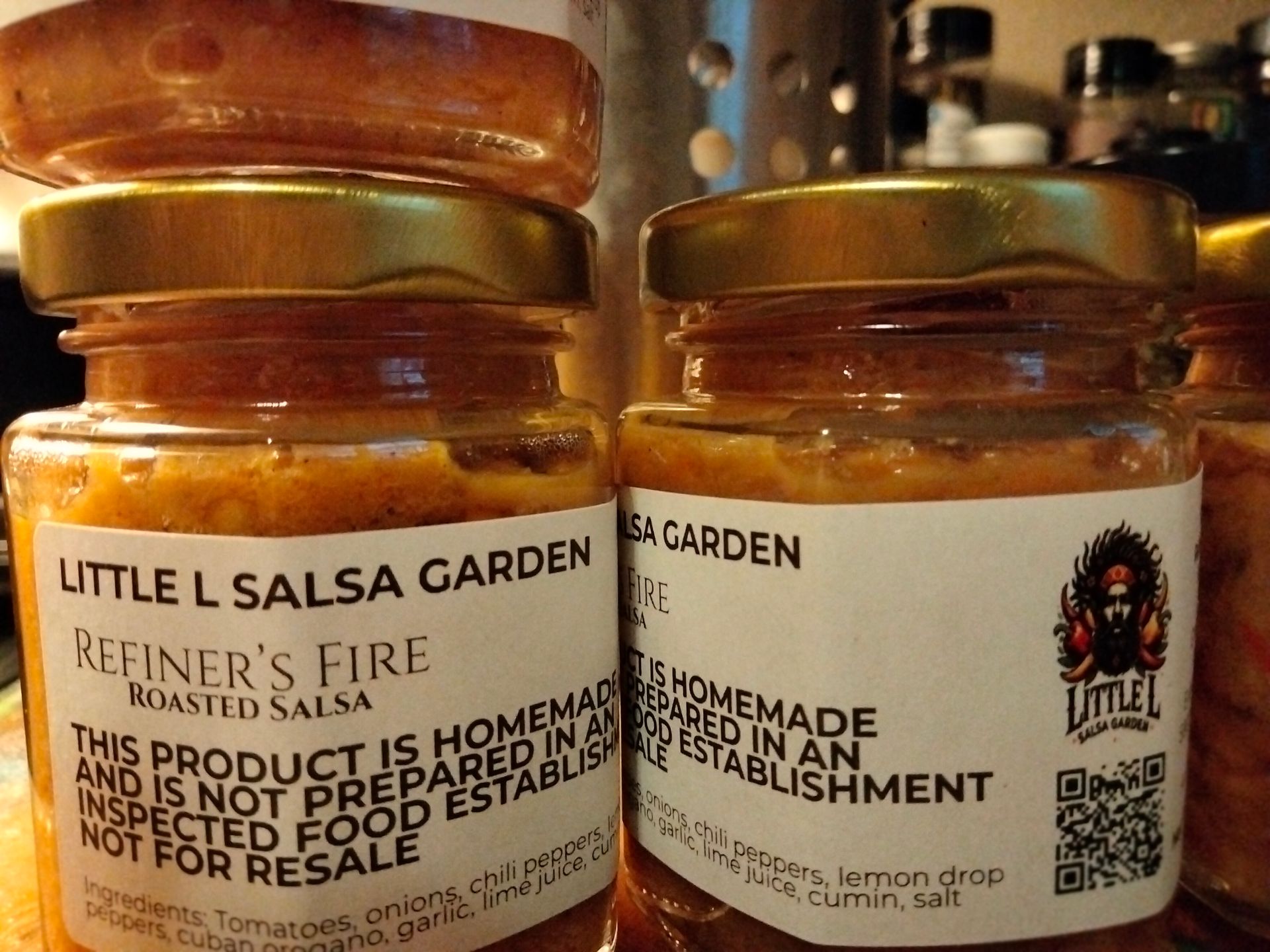 Two jars of Little L Salsa Garden roasted salsa with gold lids on a kitchen counter.