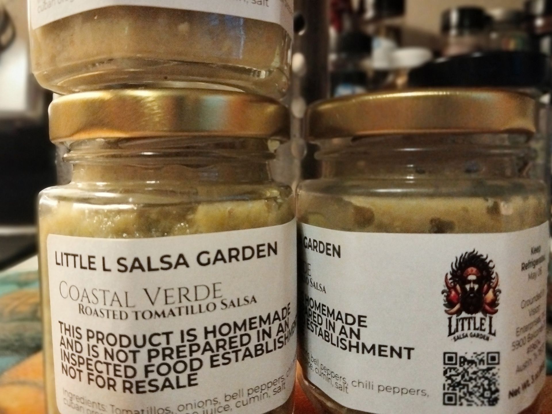 Close-up of two Little L Salsa Garden jars with labels on a shelf, one showing roasted verde tomato salsa.