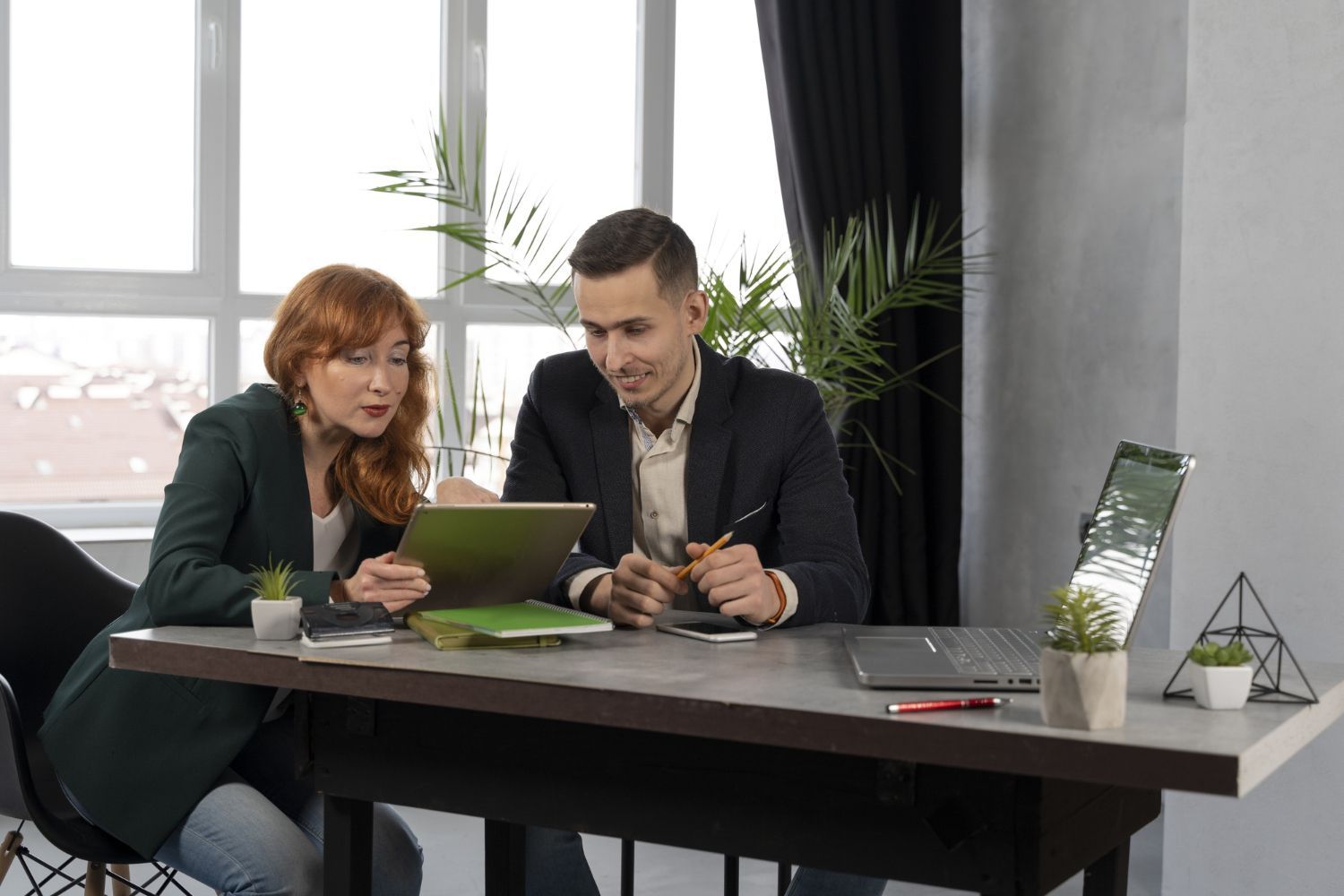 Two coworkers review a tablet at a desk in a modern office with a laptop and coffee cup nearby.