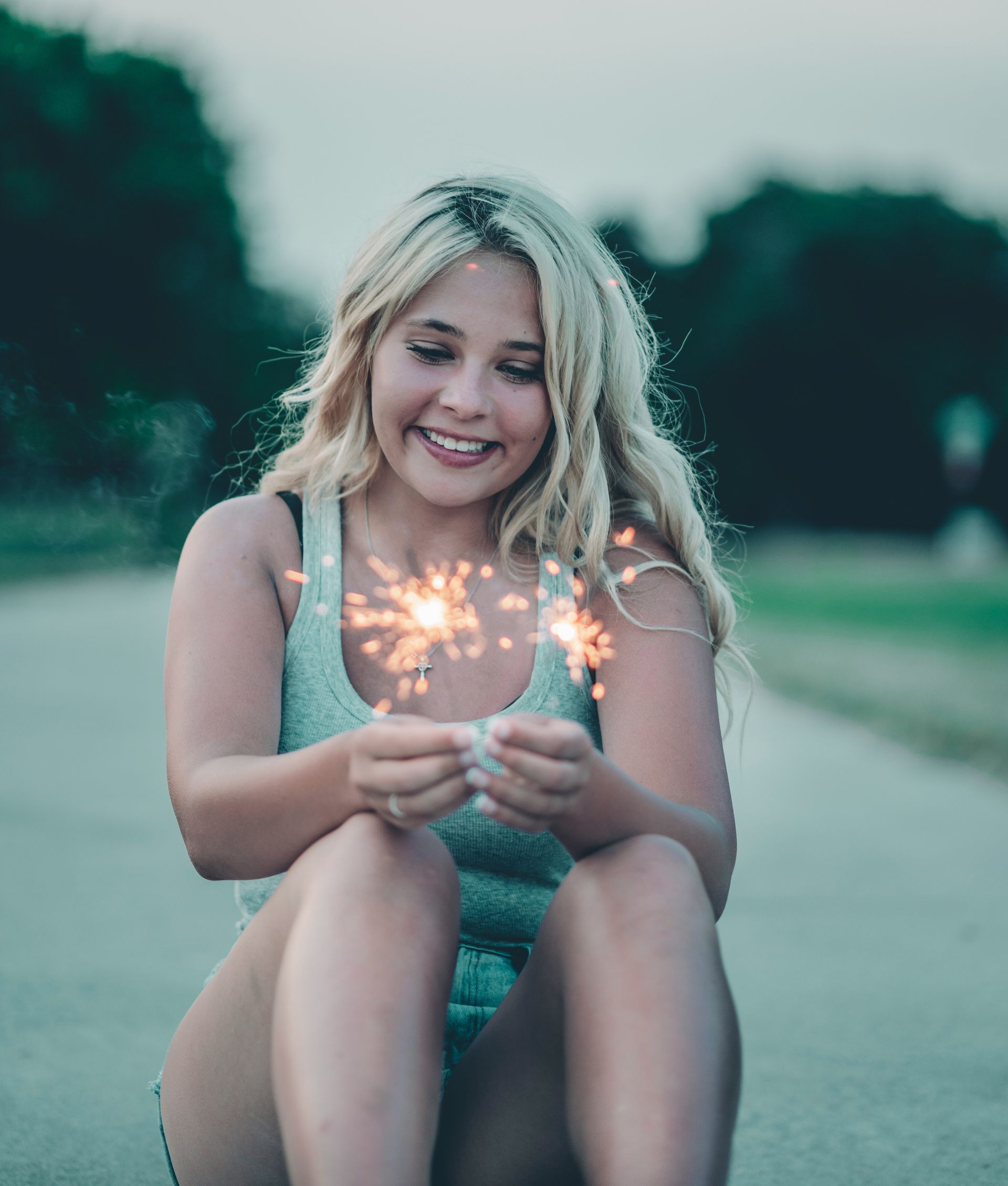 Smiling person sitting on a road holding a lit sparkler outdoors at dusk