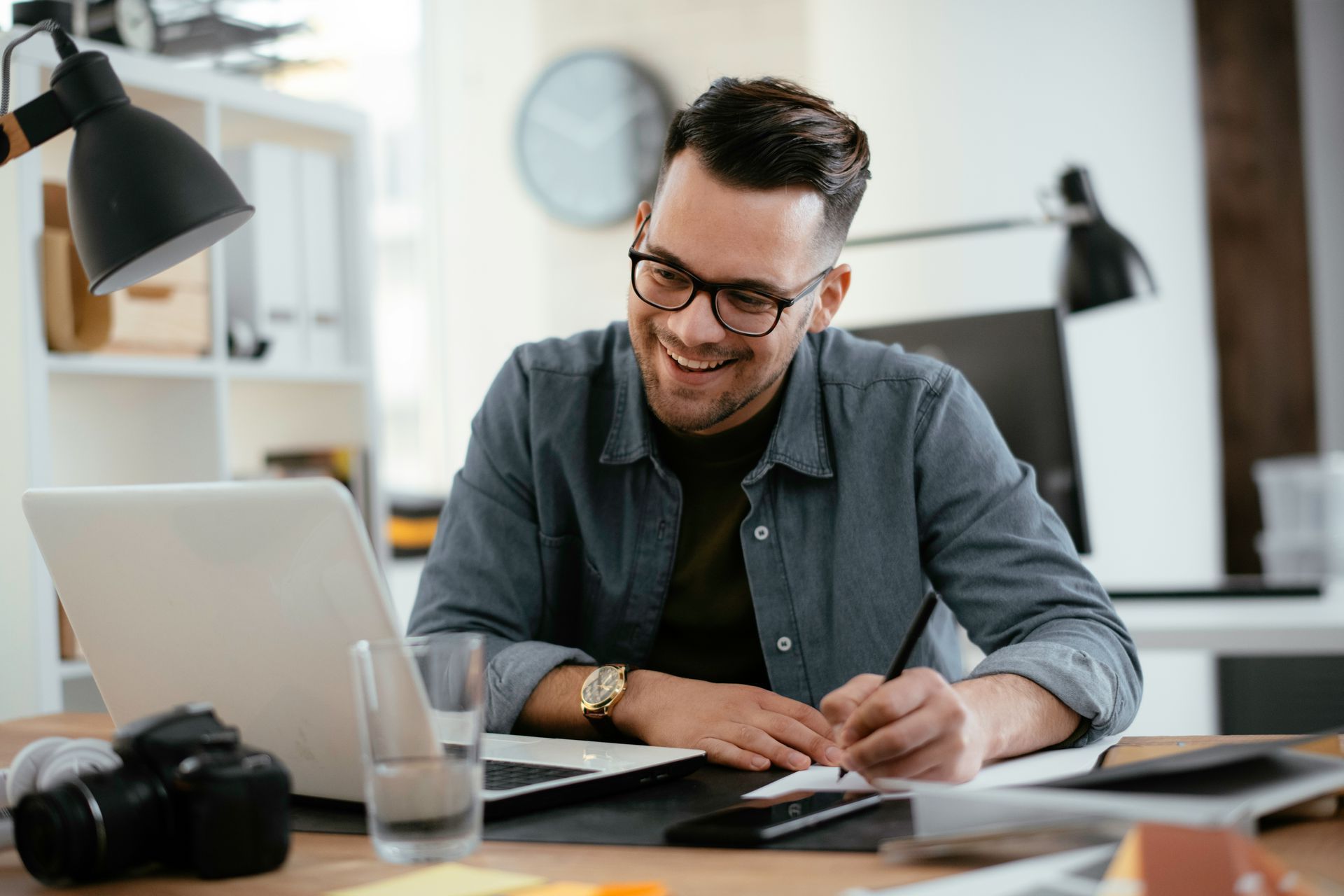 Person smiling and writing at a desk with a laptop in a bright office workspace