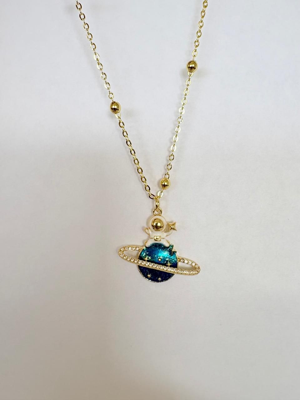 Collar de cadena dorada con un colgante azul con forma de planeta sobre fondo blanco