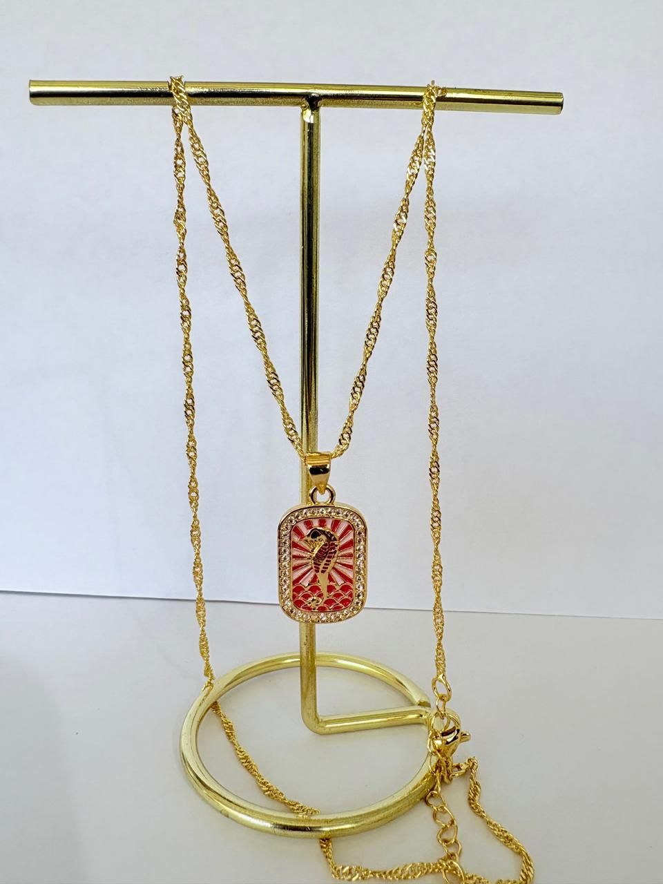 Colgante de cadena de oro colgado de un soporte dorado, con un adorno decorativo rojo y blanco.