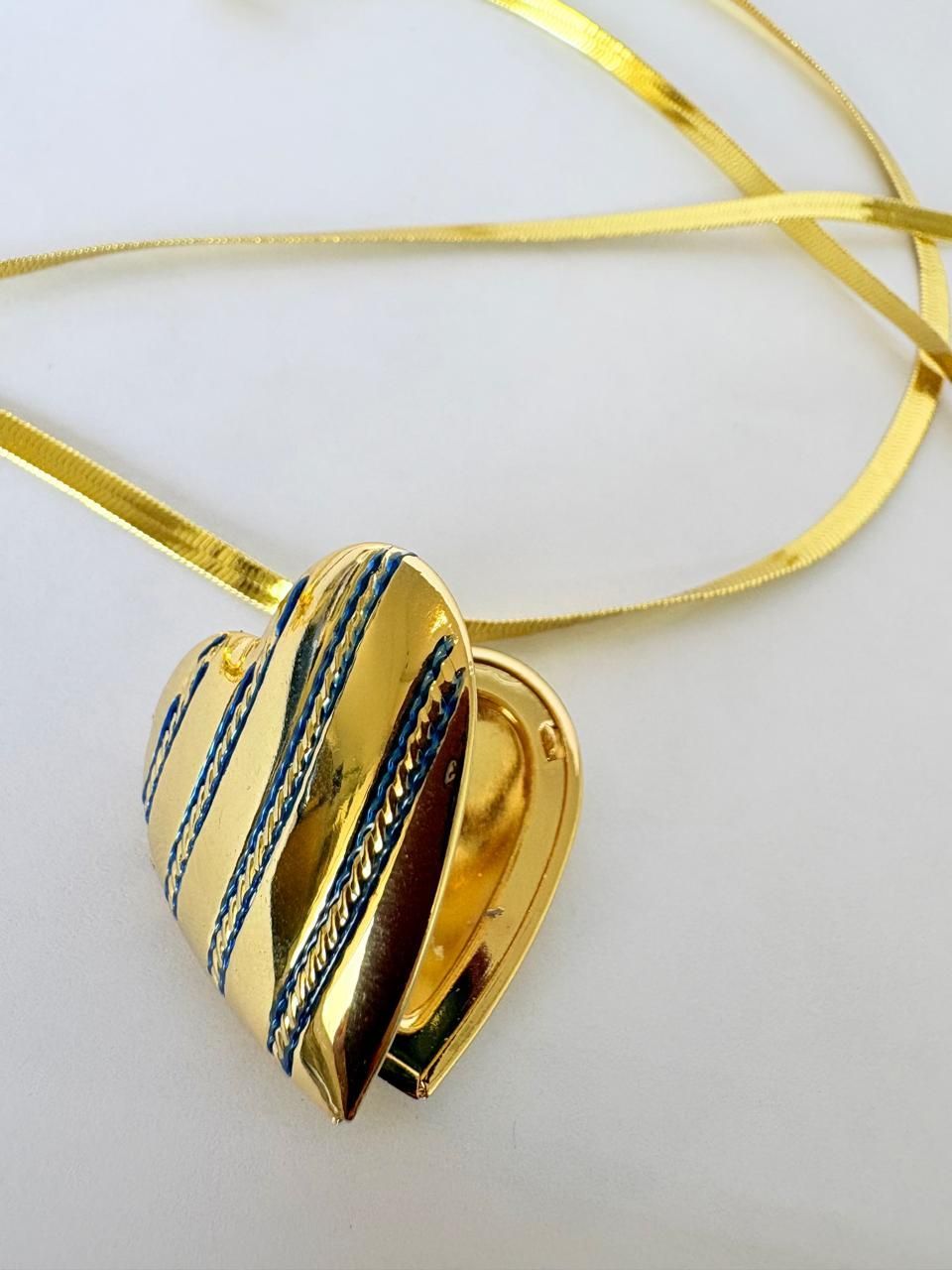 Colgante dorado en forma de corazón con franjas azules, en una fina cadena dorada, sobre un fondo claro