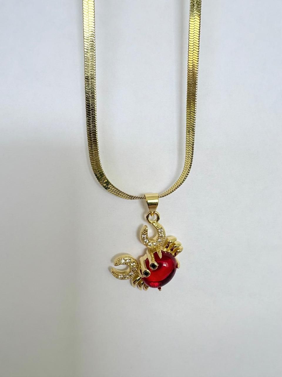 Collar dorado con un colgante de cangrejo rojo y detalles de gemas transparentes sobre un fondo claro