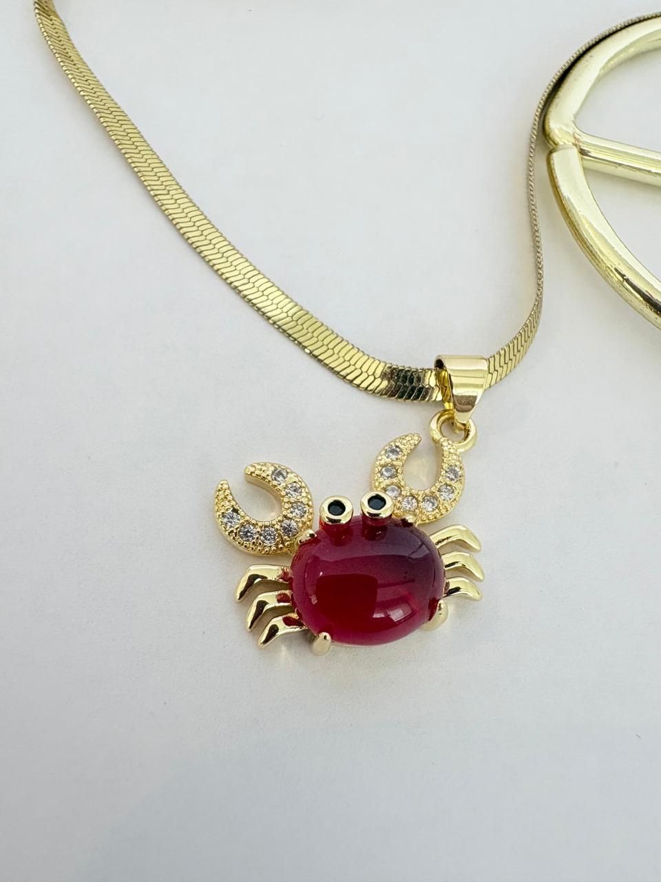 Collar dorado con un colgante de cangrejo rojo y detalles de gemas transparentes sobre un fondo claro