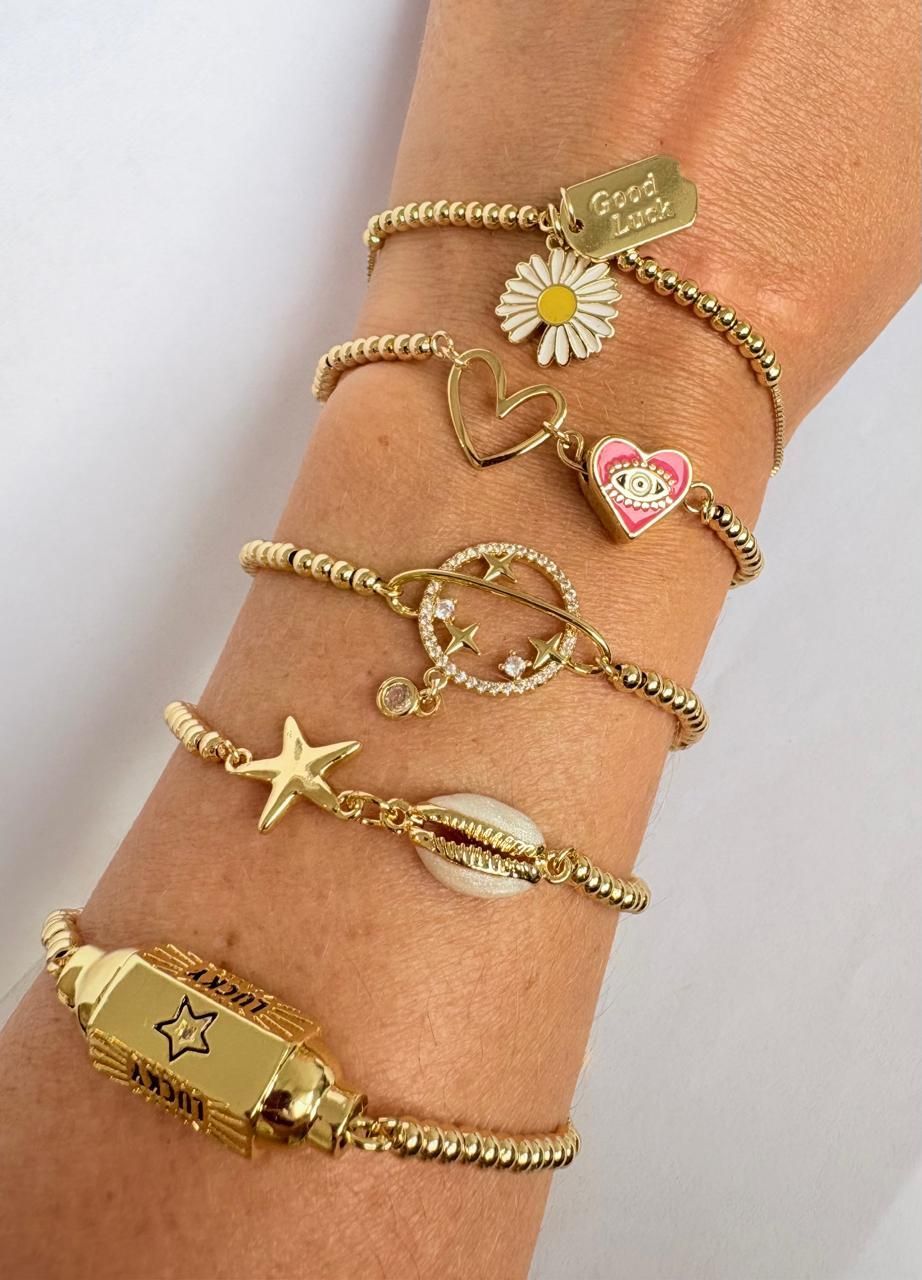 Pulseras doradas con dijes apiladas en la muñeca, con detalles de margaritas, corazones, estrellas de mar y estrellas