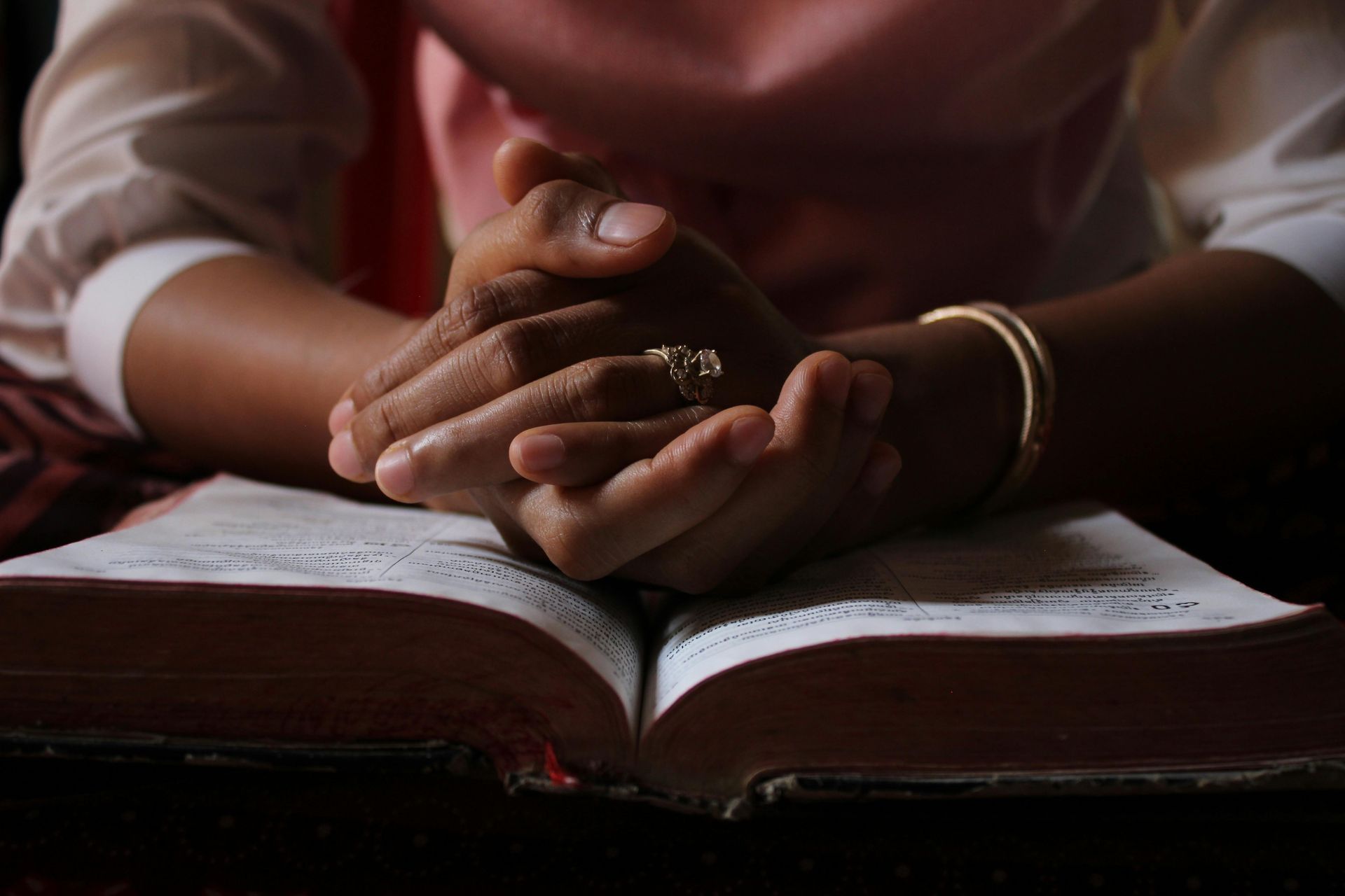 Hands clasped in prayer over an open book, with a ring and bracelet visible.