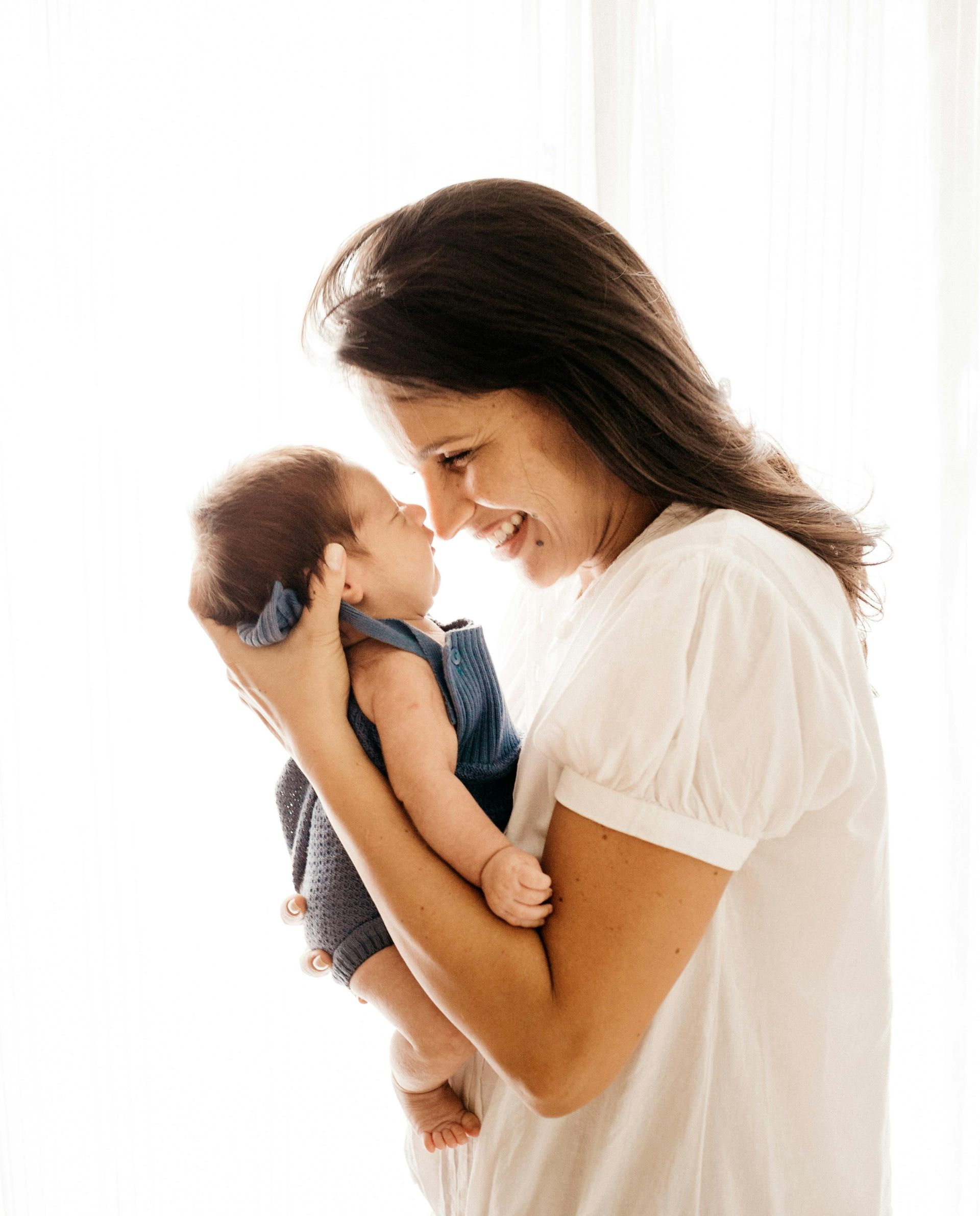Woman holding a baby, smiling and looking down. The baby is in a blue outfit. Bright, neutral background.