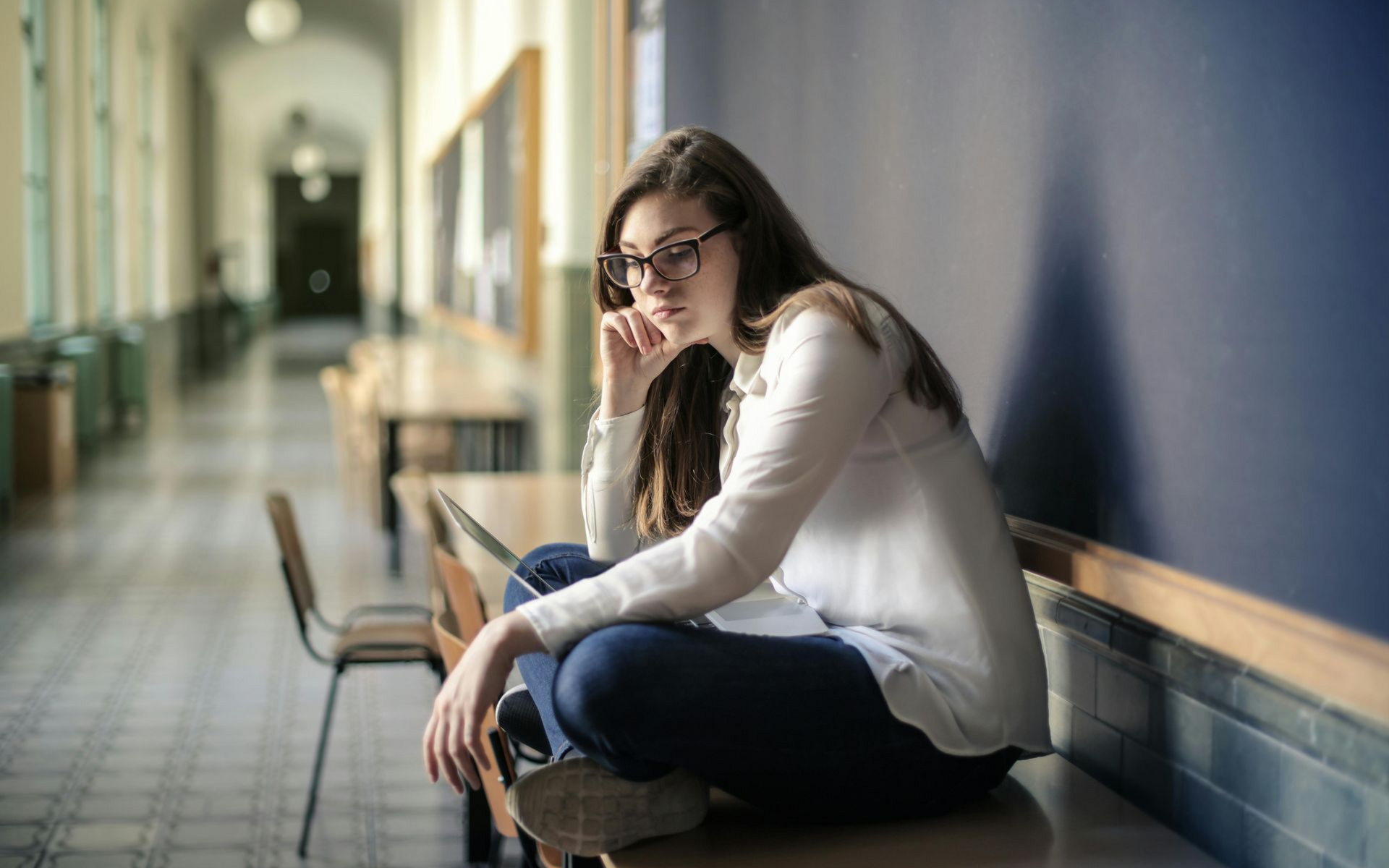 Woman with glasses sits on a table in a hallway, looking thoughtful.