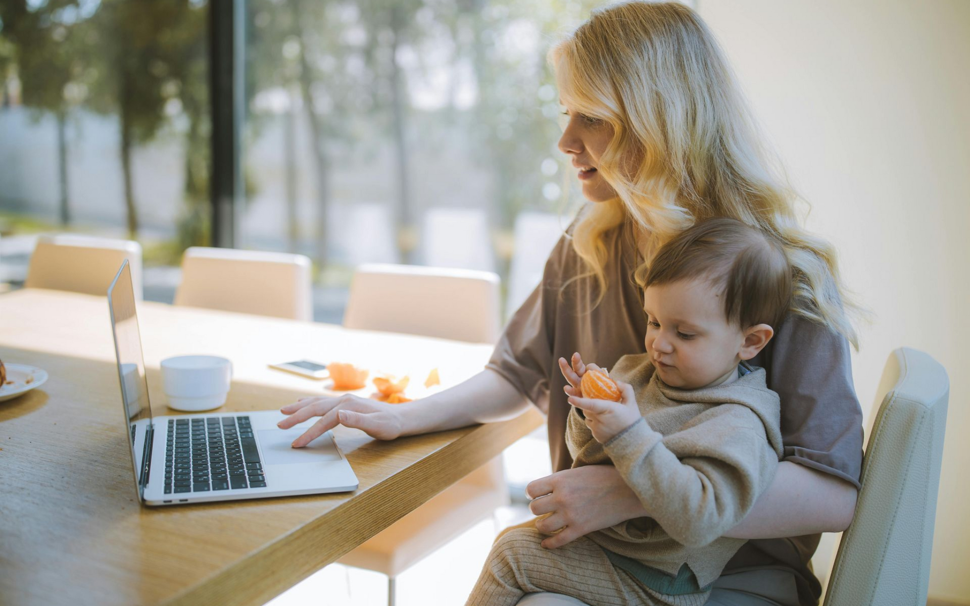 Woman working on laptop while holding a baby, seated at a table.