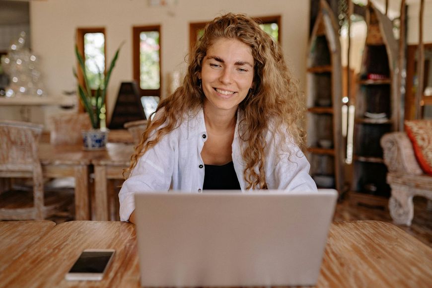Woman smiling behind a laptop at a wooden table in a cozy room