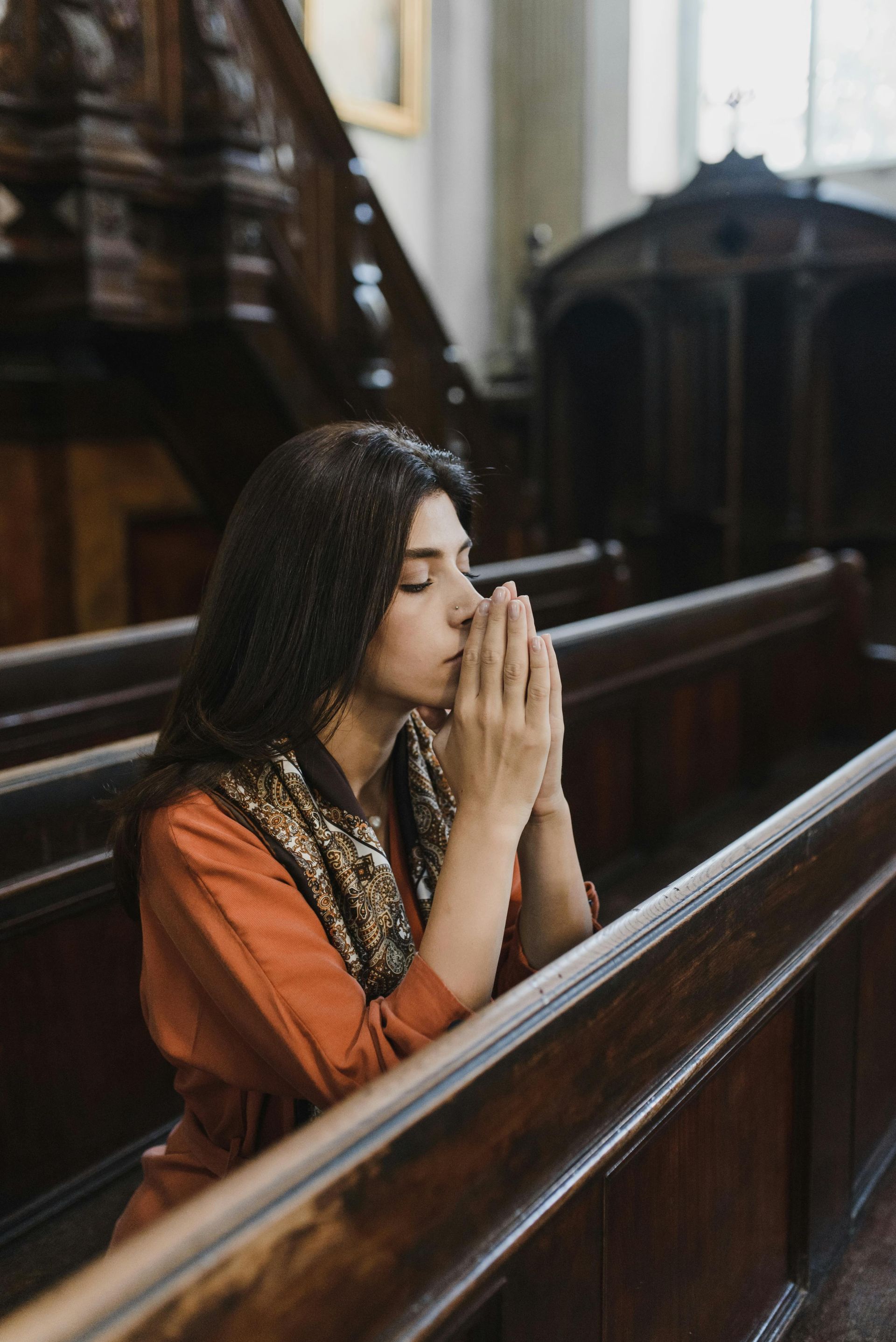 Woman praying in a wooden church pew, eyes closed, hands clasped.