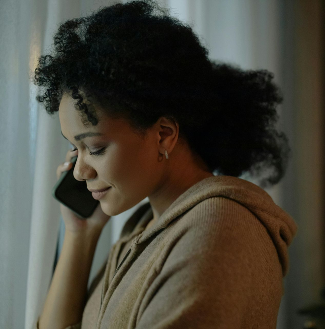 Woman in a hooded sweatshirt on a phone call near a window. She's looking down, possibly listening.