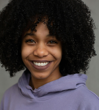 A person with curly hair smiling, wearing a purple hoodie.