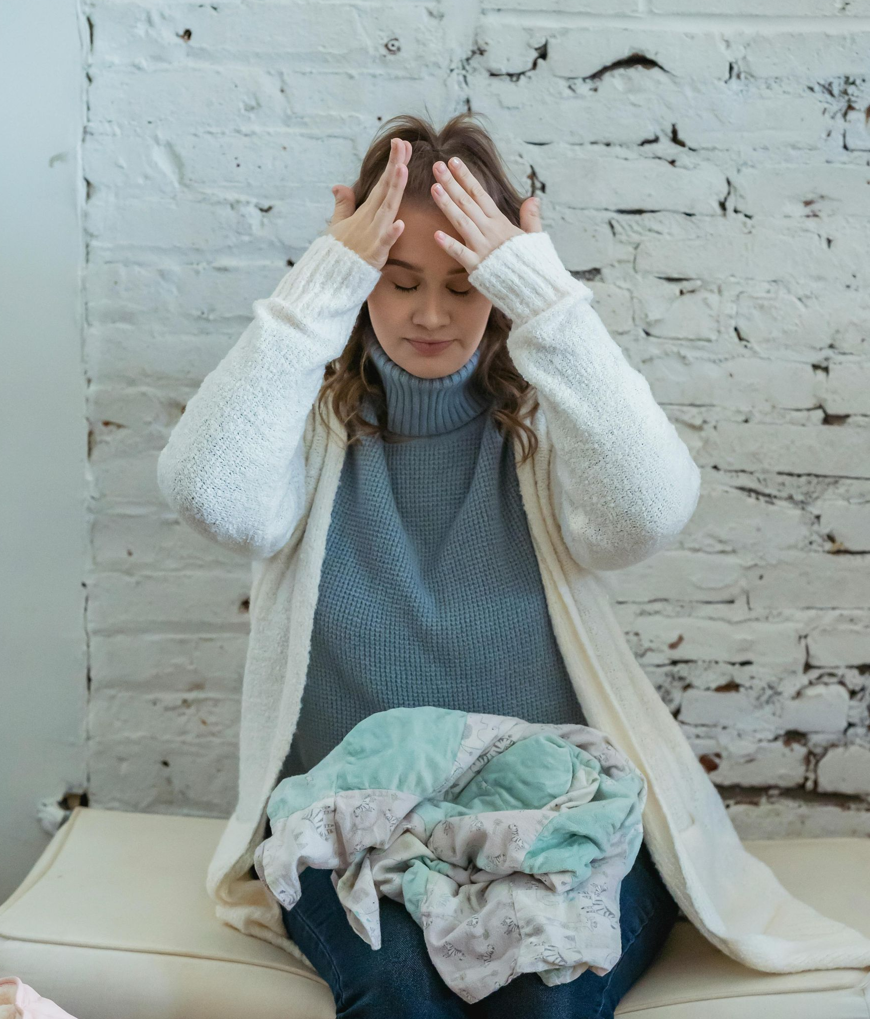 Woman with hands on head, looking down at pile of clothes, appearing stressed. White brick wall background.